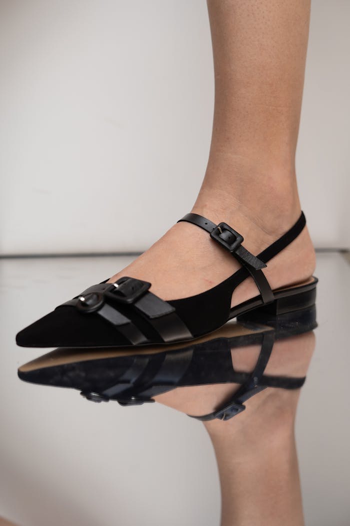 why-choose-us Close-up of a black suede slingback shoe with buckles, reflecting on a glossy surface. Fashion footwear style.
